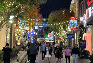 Bustling nighttime downtown promenade with trees wrapped in string lights, neon attraction signs, and crowds strolling past shops.