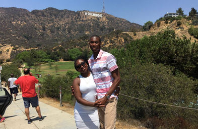 Smiling couple posing on a sunny trail in the Hollywood Hills with the iconic Hollywood sign on the hillside and green parkland below.