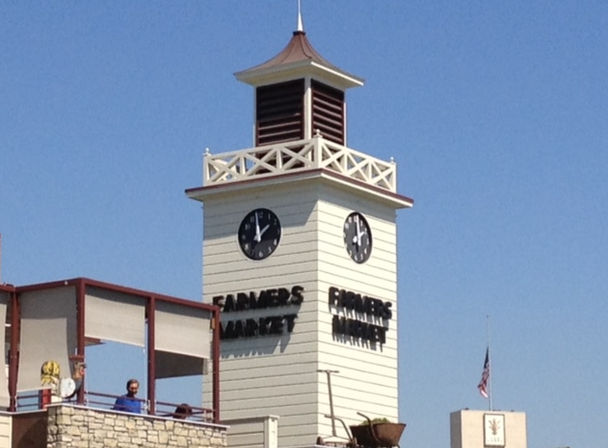 Sunny white clock tower above a farmers market building with balcony and stone facade, dual black clock faces and an American flag against a clear blue sky
