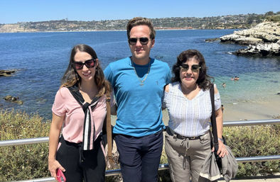 Three adults wearing sunglasses pose at an oceanfront railing on a sunny day with clear blue water, rocky shoreline and a small sandy beach in the background.