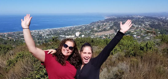 Two friends smiling and waving at a scenic hilltop overlook above a coastal town with sweeping blue ocean views