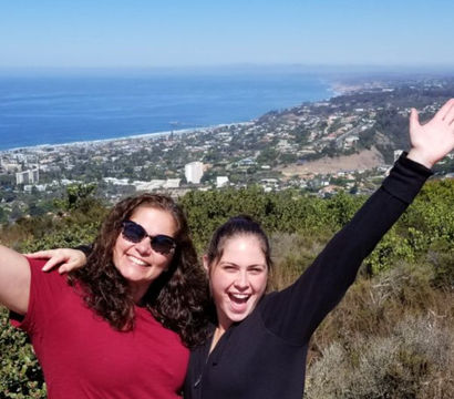 Two friends smiling and waving at a scenic hilltop overlook above a coastal town with sweeping blue ocean views