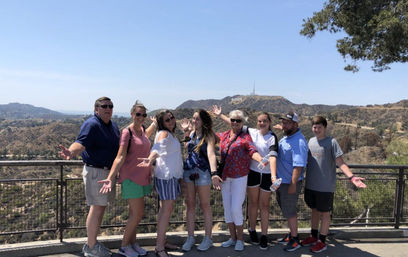 Eight vacationers (adults and teens) posing at a Hollywood Hills overlook with the Hollywood Sign visible on the distant ridge, sunny sky and rolling hills.