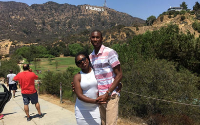 Smiling couple posing on a sunny Los Angeles hillside trail with the Hollywood Sign on the distant ridge, park greenery and other visitors in the background.
