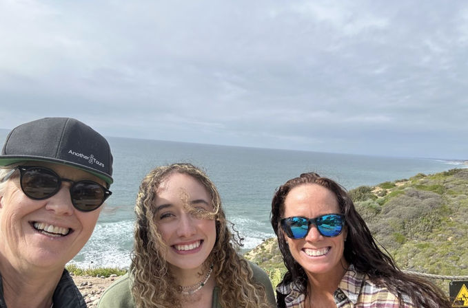 Smiling trio takes a selfie at a scenic coastal cliff overlook — sunglasses and a hat, ocean waves below, cloudy sky and coastal scrub along the rocky shoreline.