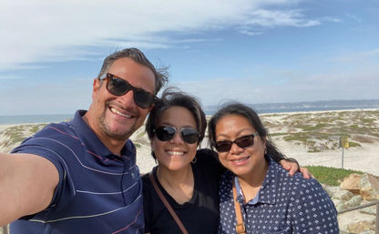 Group selfie of three friends wearing sunglasses and smiling on a sunny sandy beach with dunes and the ocean under a blue sky.