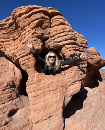 Smiling hiker popping through a natural window in layered red sandstone rock with a clear blue sky — classic Southwestern red‑rock desert scenery.