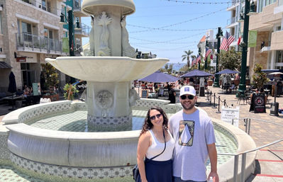 Two smiling people posing in front of an ornate stone fountain on a sunny coastal promenade with outdoor dining, string lights, American flags, palm trees and an ocean view in the background.