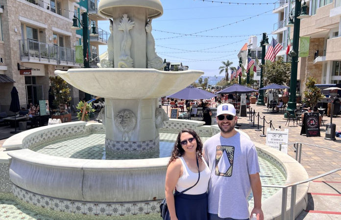 Two smiling people posing in front of an ornate stone fountain on a sunny coastal promenade with outdoor dining, string lights, American flags, palm trees and an ocean view in the background.