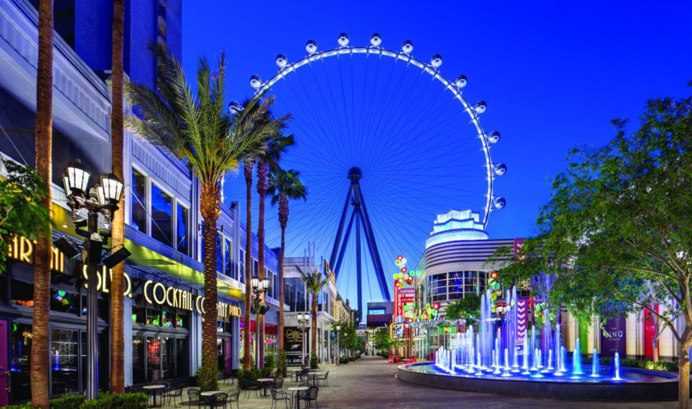 Dusk Las Vegas promenade with a giant illuminated observation wheel, palm trees, neon-lit shops and blue-lit fountain
