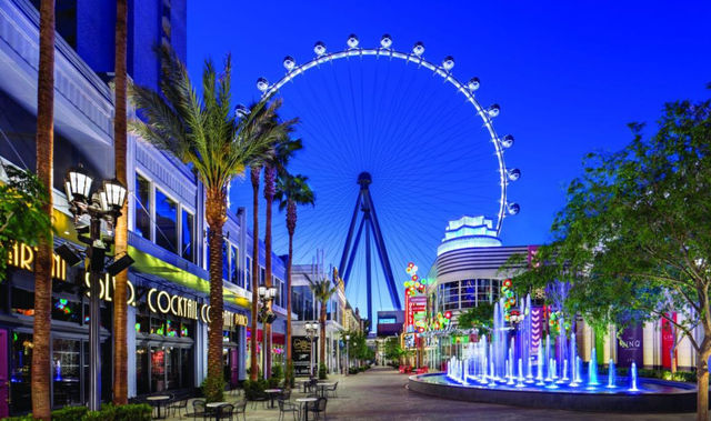 Dusk Las Vegas promenade with a giant illuminated observation wheel, palm trees, neon-lit shops and blue-lit fountain