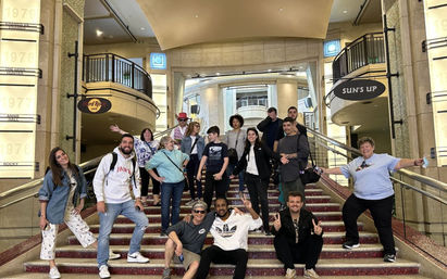 About 16 people smiling and posing on a wide, carpeted grand staircase inside an ornate multi-level lobby with balconies, glass railings and illuminated wall panels — casual group photo in a theater or hotel-style interior.