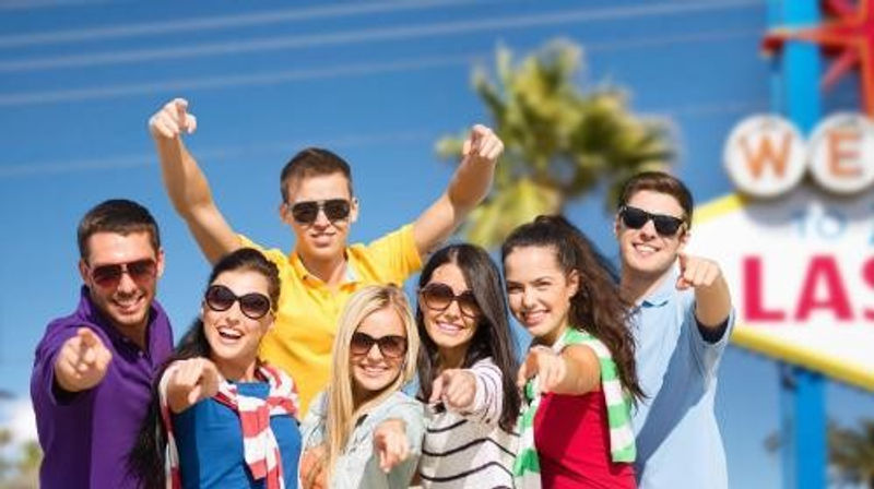 Smiling group of young friends pointing at the camera in a sunny outdoor tourist spot with palm trees and a colorful sign, vacation vibe