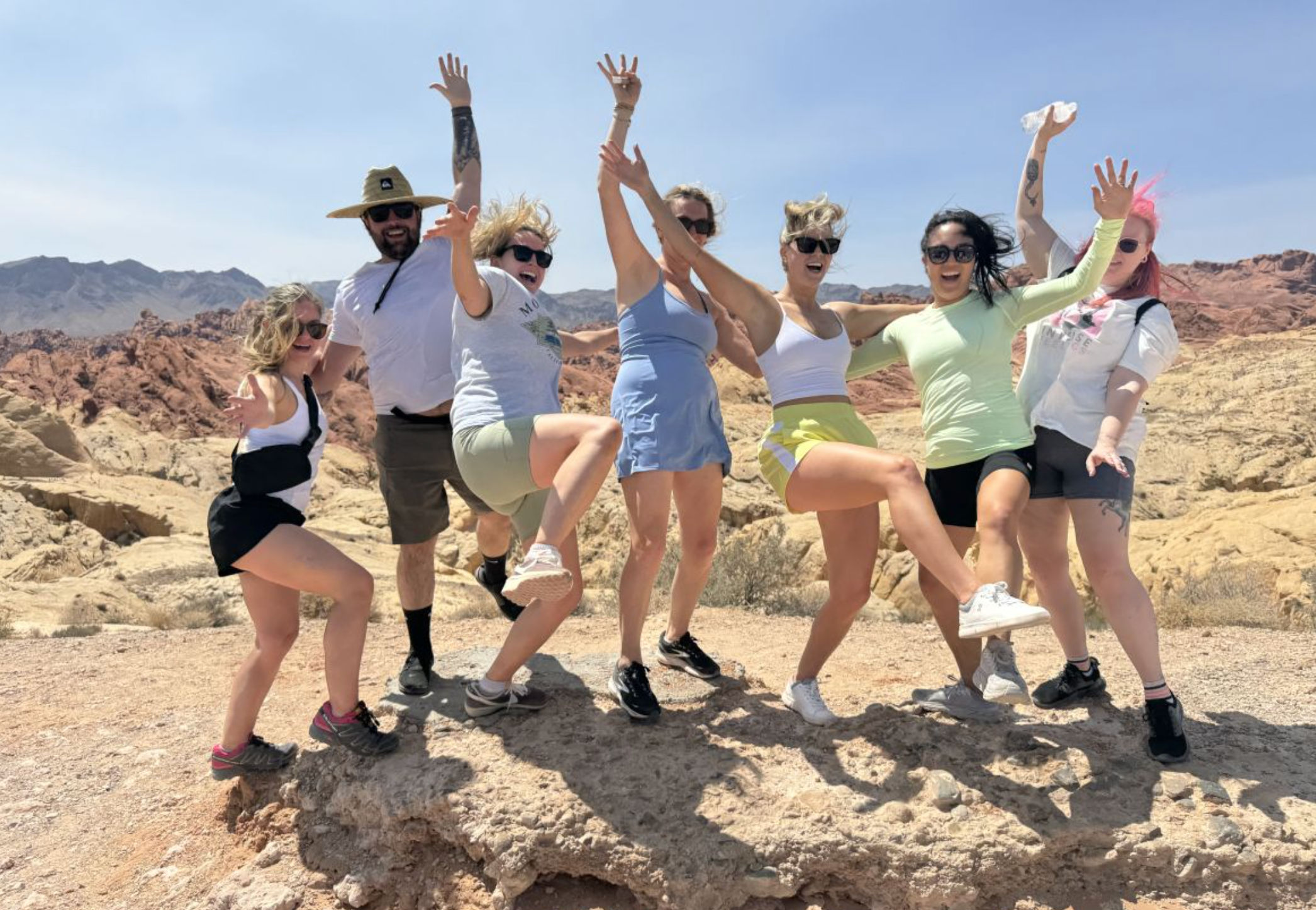 Seven friends in bright activewear jump and pose on a sunlit red‑rock desert outcrop — hiking group photo, outdoor adventure in a rocky desert landscape.