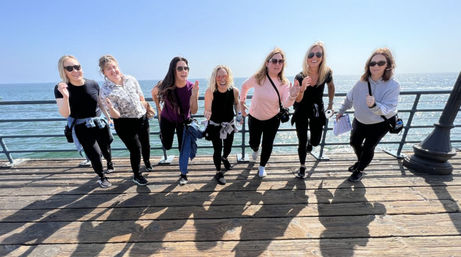 Seven women pretending to run toward the camera on a sunny oceanfront pier, smiling and casting long shadows on weathered wooden planks.