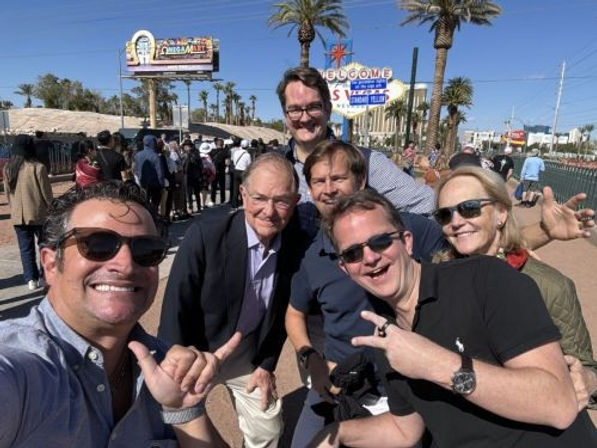 Cheerful group selfie by the iconic 'Welcome to Las Vegas' sign on a sunny, palm-lined sidewalk with tourists and billboards in the background.