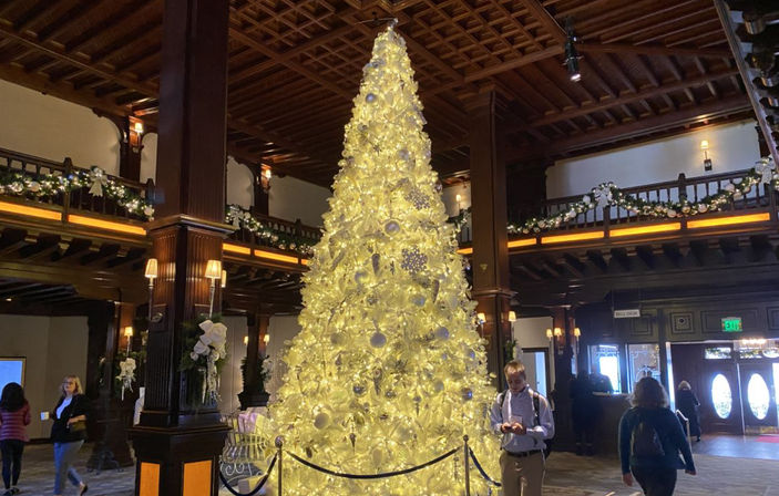 Tall glowing white-and-gold Christmas tree in a grand wood-paneled hotel lobby with garlanded balconies, coffered ceiling, roped-off base, and people walking nearby.