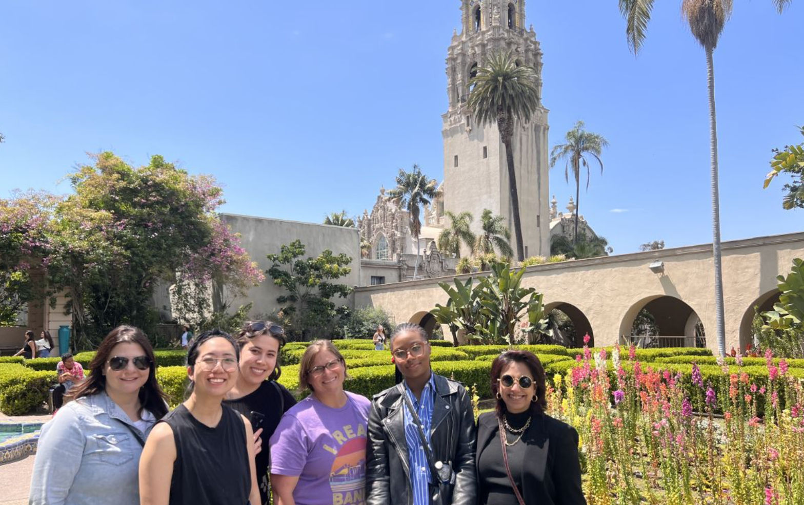 Group of six adults smiling in a sunlit botanical garden with colorful flower beds, arched colonnade and historic Spanish‑style tower rising behind — Balboa Park, San Diego.