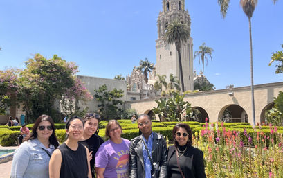 Group of six adults smiling in a sunlit botanical garden with colorful flower beds, arched colonnade and historic Spanish‑style tower rising behind — Balboa Park, San Diego.