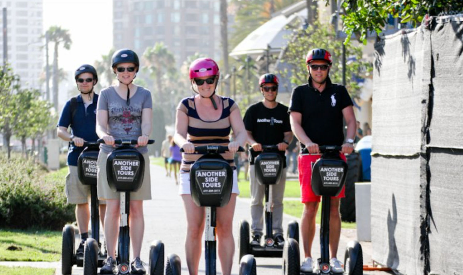 Five adults wearing helmets on a Segway tour along a sunny beachside promenade lined with palm trees and high-rise buildings.