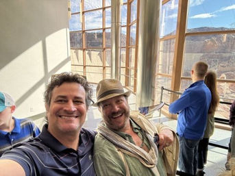 Two smiling men take a selfie inside a sunlit visitor center with tall glass windows overlooking a rocky canyon, while other visitors gaze at the scenic desert cliffs.
