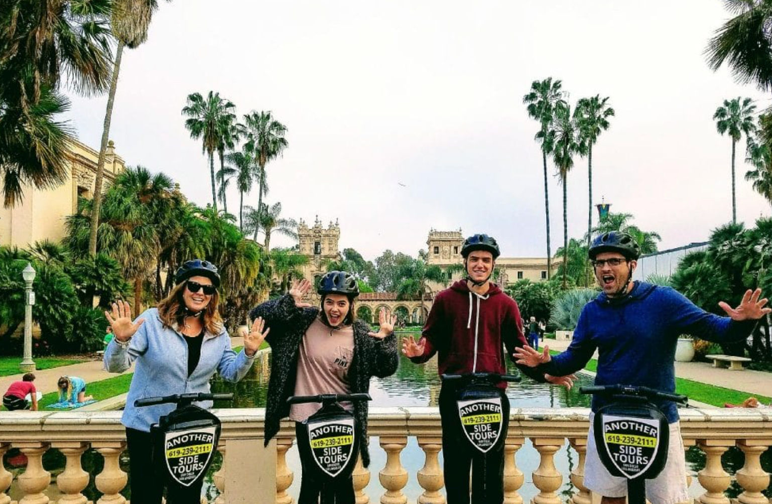 Four people on a guided electric scooter tour at Balboa Park, San Diego, posing by a reflecting pool with palm trees and historic Spanish-style buildings.