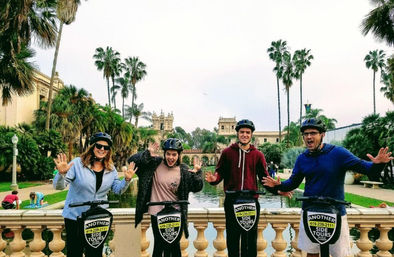 Four people on a guided electric scooter tour at Balboa Park, San Diego, posing by a reflecting pool with palm trees and historic Spanish-style buildings.