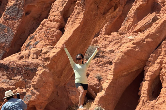 Person in sunglasses and activewear poses with arms raised on red sandstone rock formations while another person in a hat photographs the sunny desert canyon scene