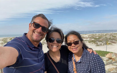 Beach selfie of three smiling friends wearing sunglasses with sandy dunes and the ocean shoreline in the background