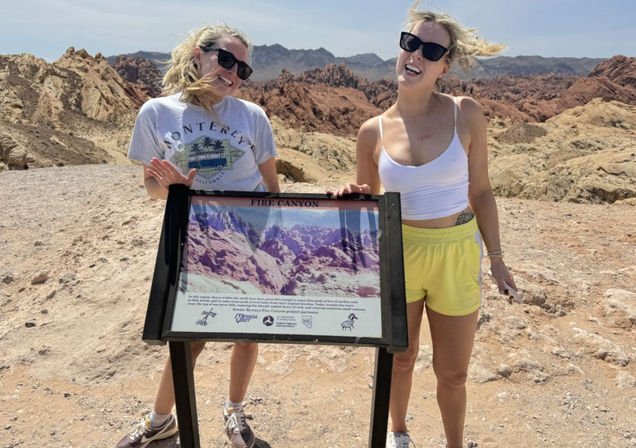 Two smiling women in sunglasses pose by a 'Fire Canyon' interpretive sign among red and tan sandstone formations at Valley of Fire State Park, Nevada on a sunny day
