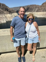 Two adults in light summer clothes smiling and posing at a concrete dam overlook with deep-blue reservoir and arid canyon cliffs under a clear sky.