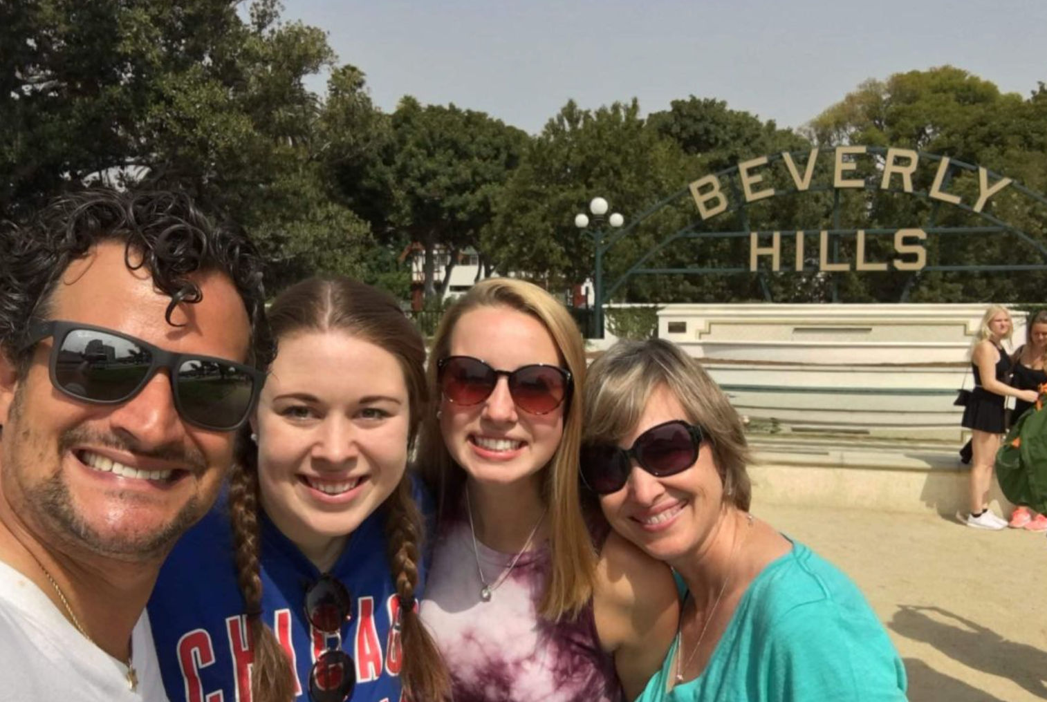 Smiling group selfie of four adults in casual summer clothes, two wearing sunglasses, posed in front of the iconic "Beverly Hills" sign and fountain in sunny Beverly Hills, California.