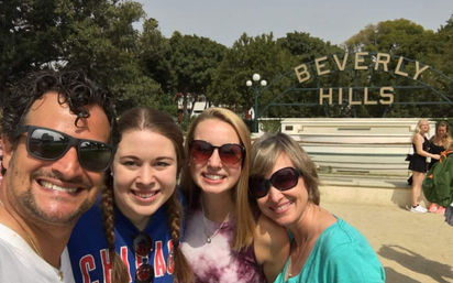 Selfie of four smiling adults in sunglasses and casual clothes in front of the Beverly Hills sign and fountain on a sunny day
