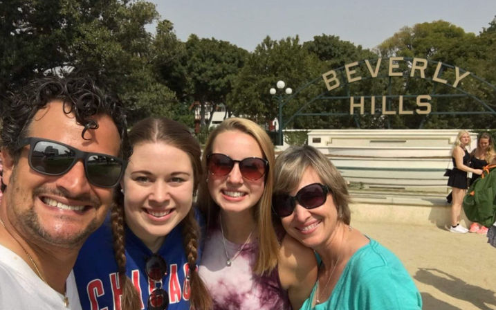 Selfie of four smiling adults in sunglasses and casual clothes in front of the Beverly Hills sign and fountain on a sunny day