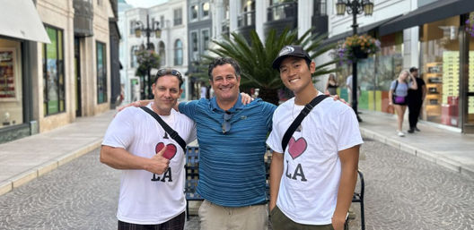 Three smiling men posing arm-in-arm on a cobblestone pedestrian shopping street, two wearing white “I ♥ LA” shirts and one in a blue striped polo with palm plant and storefronts in the background — Los Angeles outdoor shopping area.