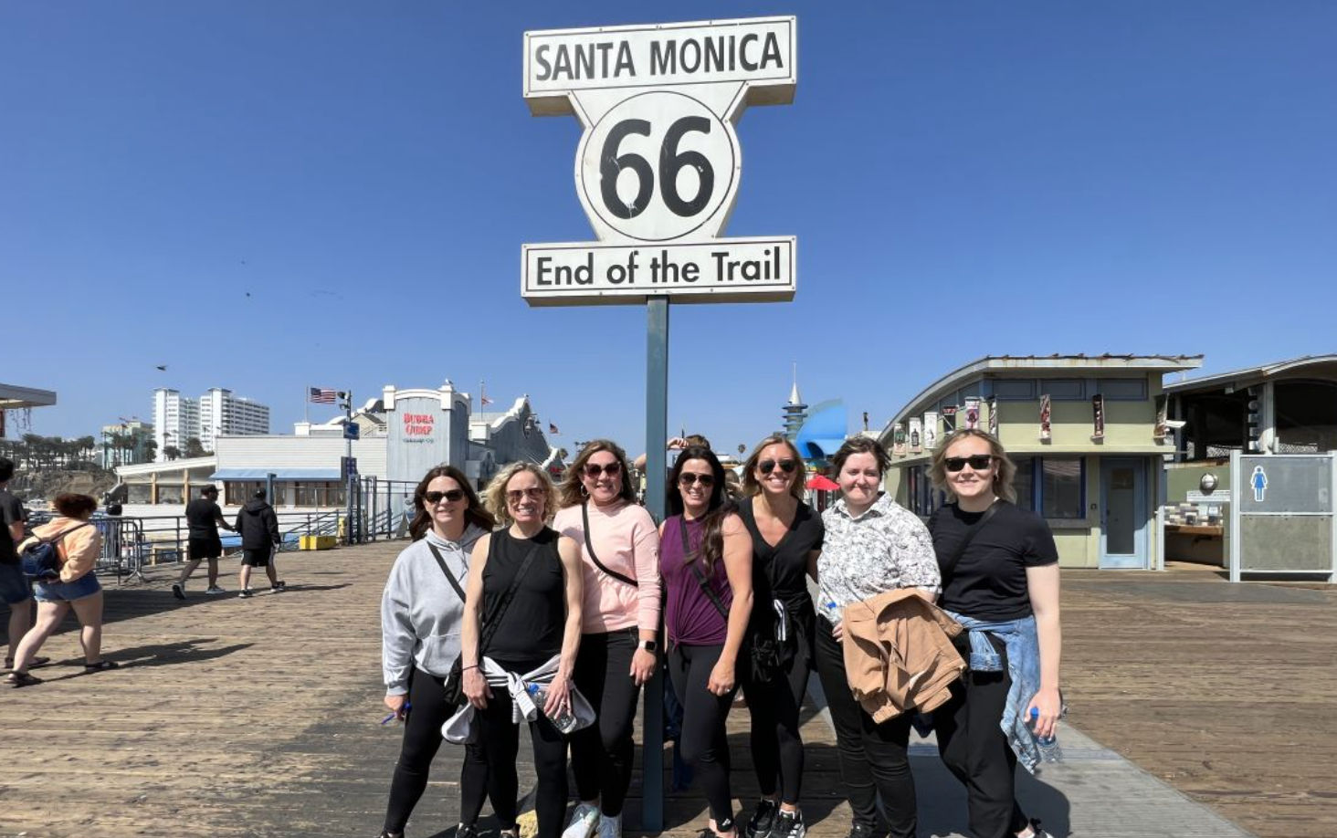 Group of eight women smiling under the Route 66 “Santa Monica – End of the Trail” sign on the Santa Monica Pier boardwalk, bright blue sky and pier attractions in the background.
