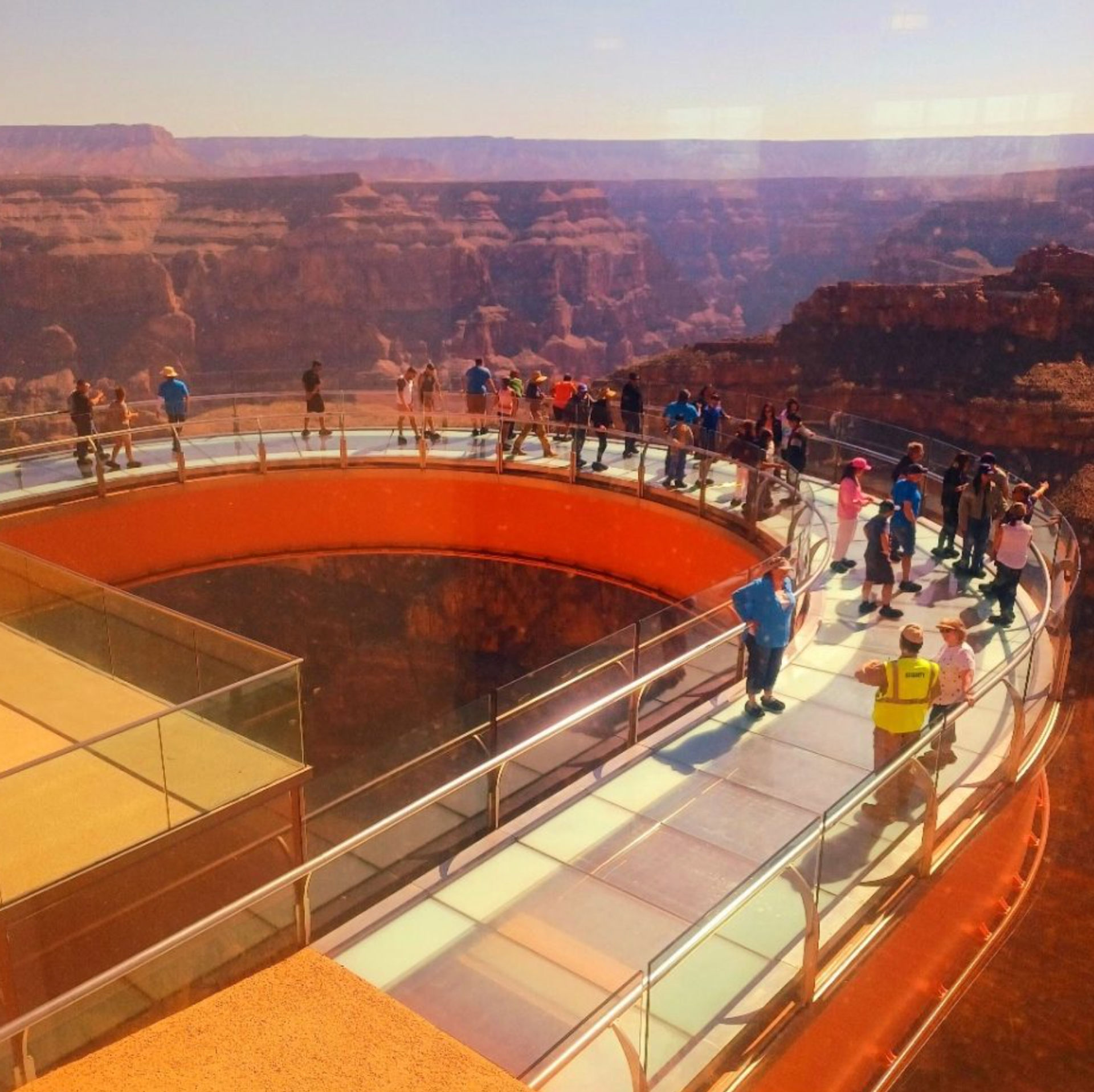 Curved glass‑bottom observation deck jutting over a dramatic red‑rock desert canyon, visitors lining the railing to enjoy panoramic cliffside views under a clear sky