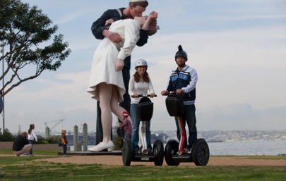 Two people in helmets ride Segways on a grassy waterfront path, smiling in front of a giant public sculpture of a sailor kissing a nurse, with onlookers and a distant city skyline behind them.