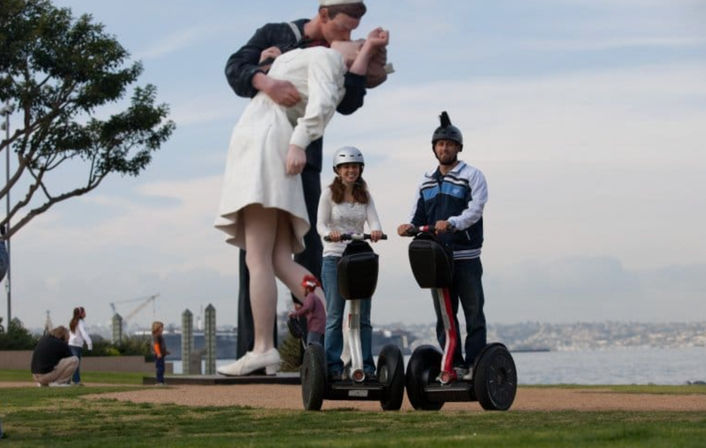Two people in helmets ride Segways on a grassy waterfront path, smiling in front of a giant public sculpture of a sailor kissing a nurse, with onlookers and a distant city skyline behind them.