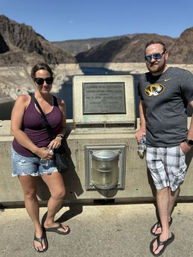 Two tourists in sunglasses lean on a concrete dam railing beside a commemorative plaque, overlooking a deep-blue reservoir and rugged desert canyon under a clear sky.