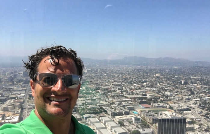 Smiling man in sunglasses takes a rooftop selfie overlooking a sprawling Los Angeles cityscape and distant hills on a clear sunny day.