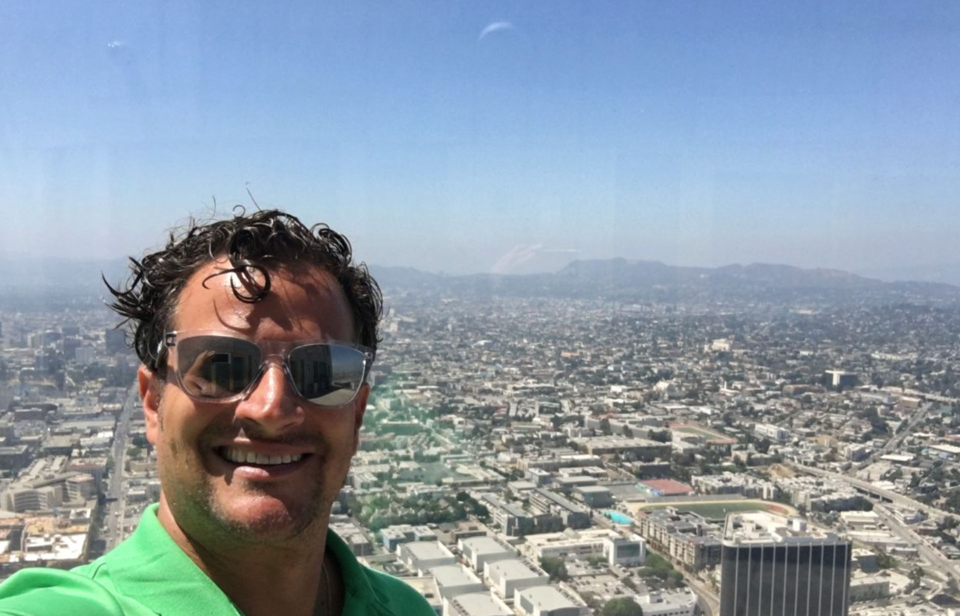Smiling man in sunglasses takes a rooftop selfie overlooking a sprawling Los Angeles cityscape and distant hills on a clear sunny day.