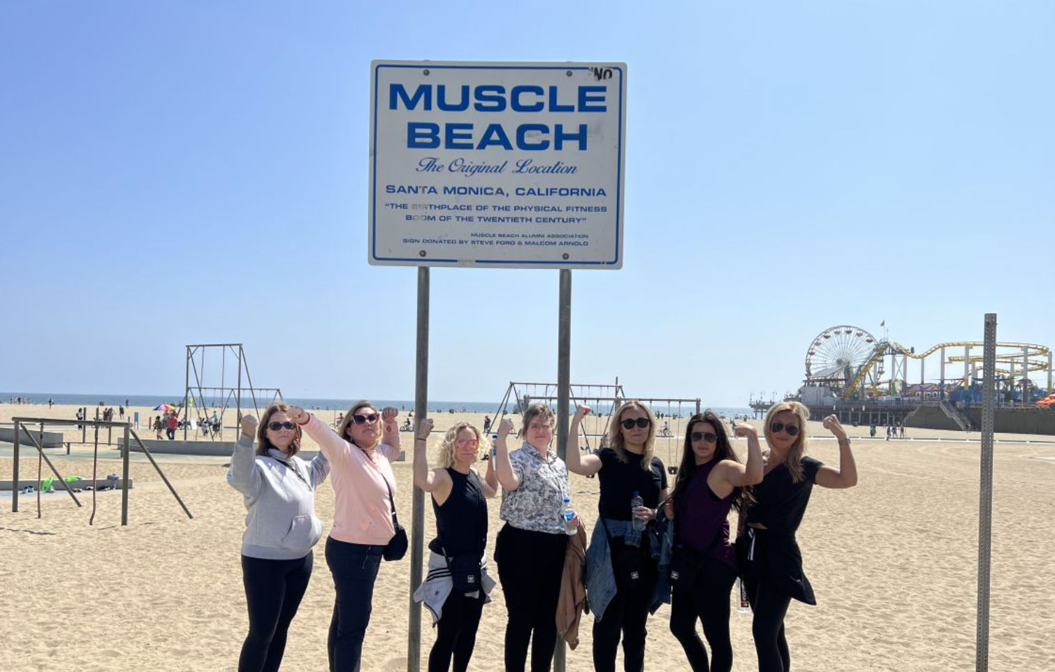 Seven women flexing and posing under a vintage beach fitness sign on a sunny Santa Monica shoreline with an amusement pier and Ferris wheel in the background.