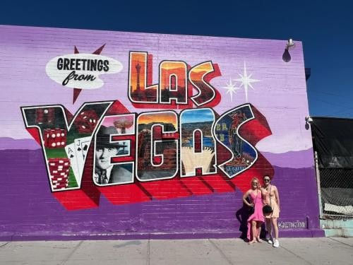 Colorful postcard-style mural on a purple brick wall reading "Greetings from LAS VEGAS" with dice, playing cards and skyline images; two people pose on the sidewalk in front under a clear blue sky.