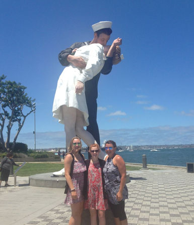 Giant sculpture of a sailor kissing a nurse on the San Diego waterfront promenade, three women posing at its base with blue sky, harbor and sailboats in the background.