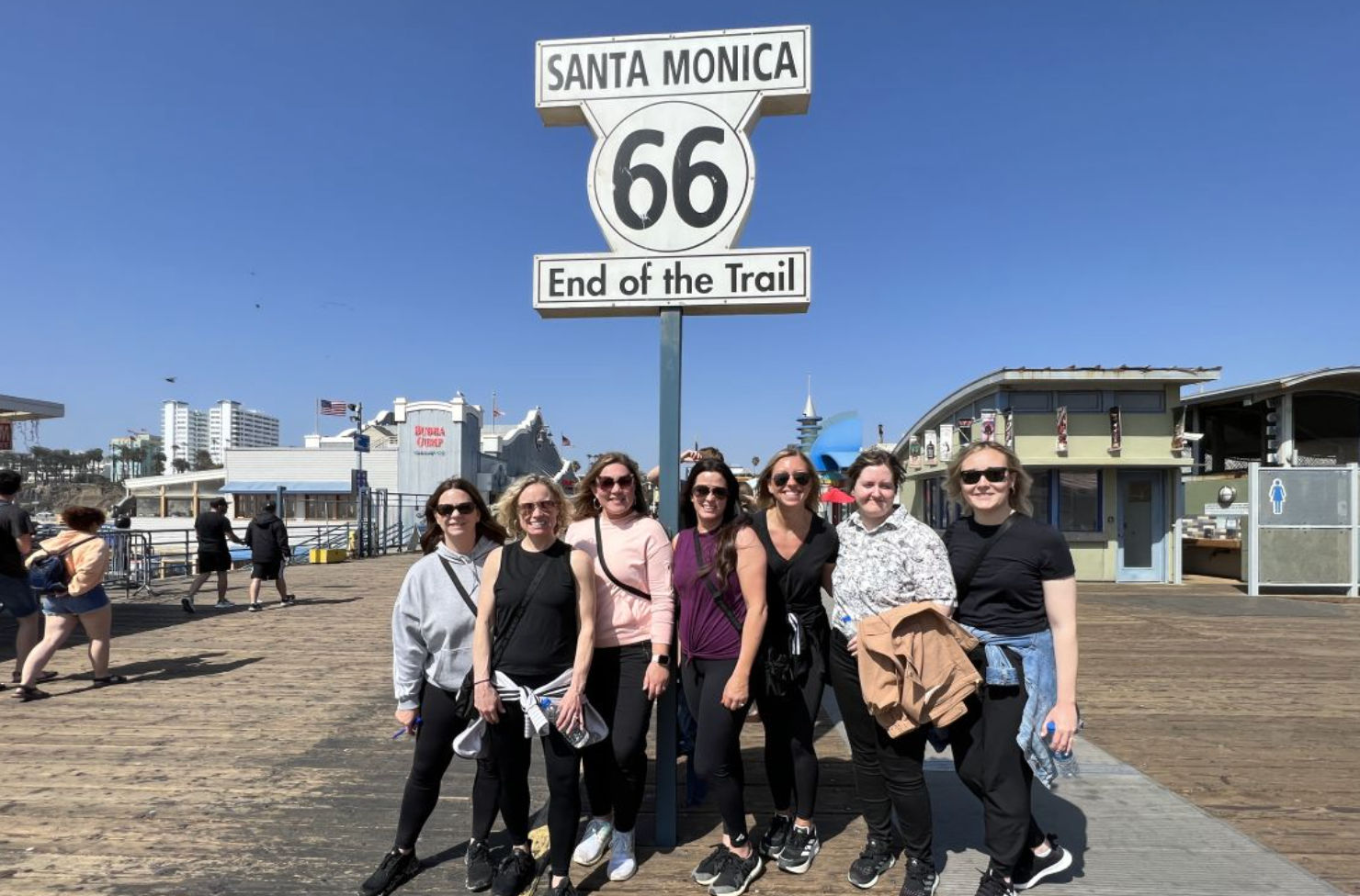 Smiling group of women posing under the iconic Santa Monica Route 66 "End of the Trail" sign on a sunny Santa Monica Pier boardwalk with oceanfront buildings and clear blue sky.