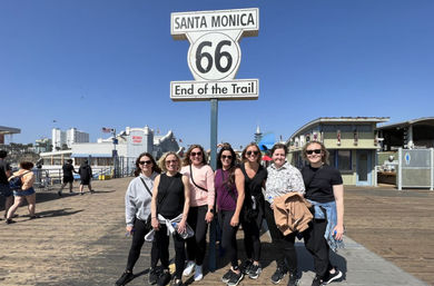Smiling group of women posing under the iconic Santa Monica Route 66 "End of the Trail" sign on a sunny Santa Monica Pier boardwalk with oceanfront buildings and clear blue sky.