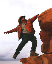 Person in a cowboy hat and orange leather jacket reaching between red sandstone boulders on a cloudy day, striking an adventurous cliffside pose in a southwestern red-rock landscape.