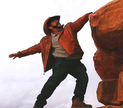 Person in a cowboy hat and orange leather jacket reaching between red sandstone boulders on a cloudy day, striking an adventurous cliffside pose in a southwestern red-rock landscape.