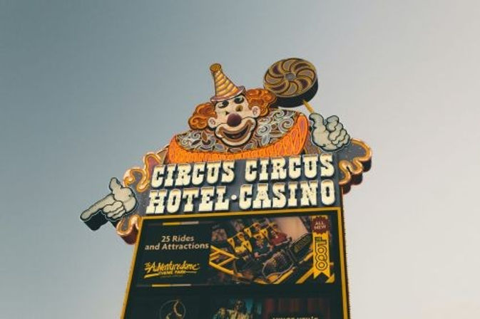 Colorful retro clown marquee advertising a hotel-casino, featuring a pointing clown with a swirl lollipop and party hat above a poster for carnival rides against a pale sky.
